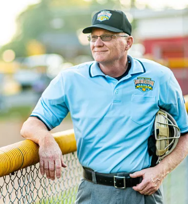 Man holding an umpire mask and resting his arm on a fence surrounding a baseball field